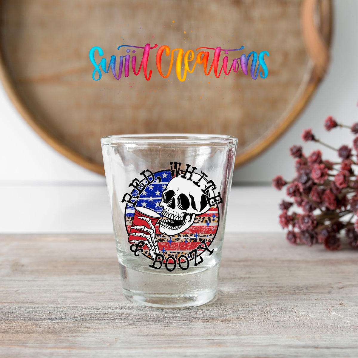 Red White and Boozy shot glass transfer on a clear glass beside a wooden barrel and dried flowers
