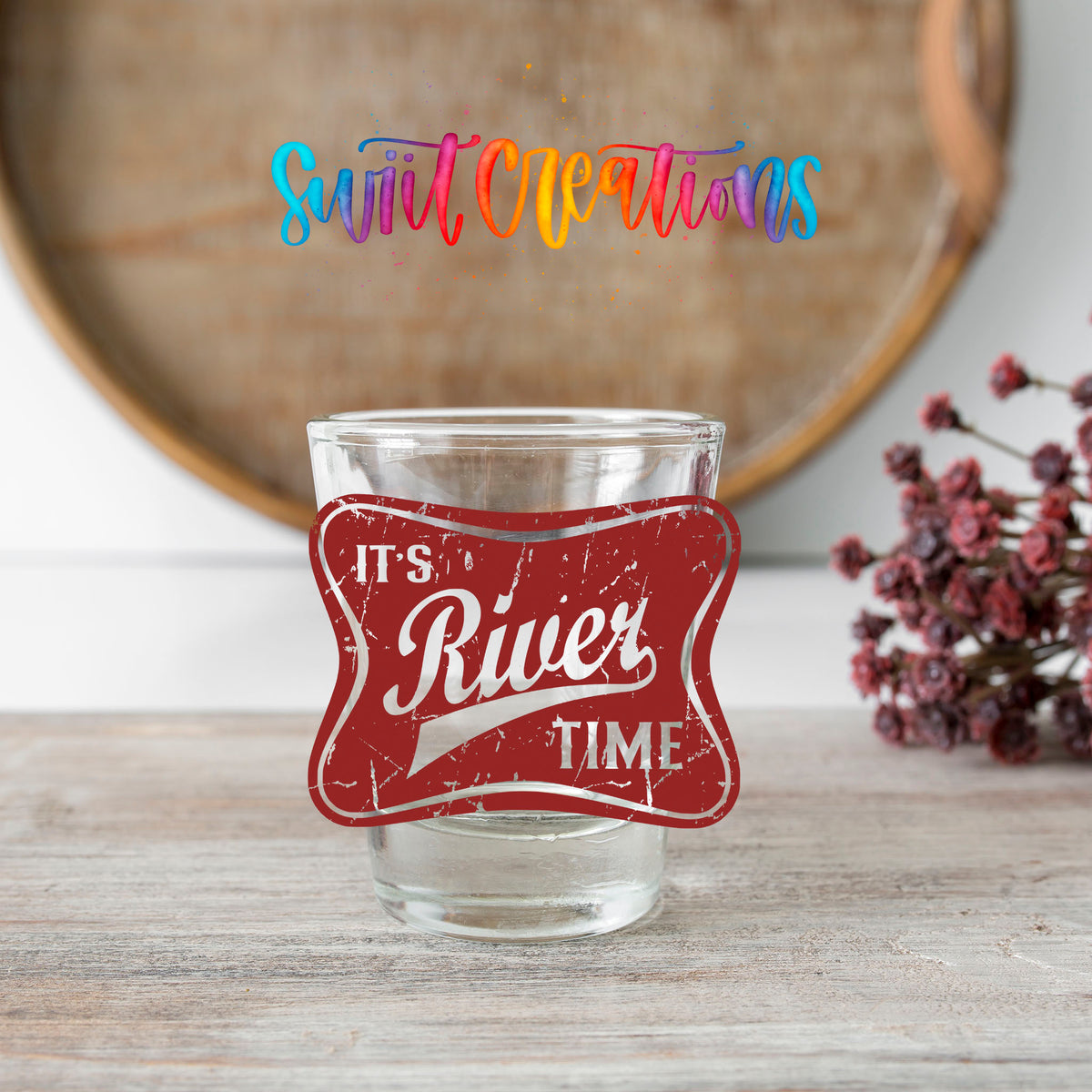 River Time shot glass transfer on a clear glass beside a wooden barrel and dried flowers