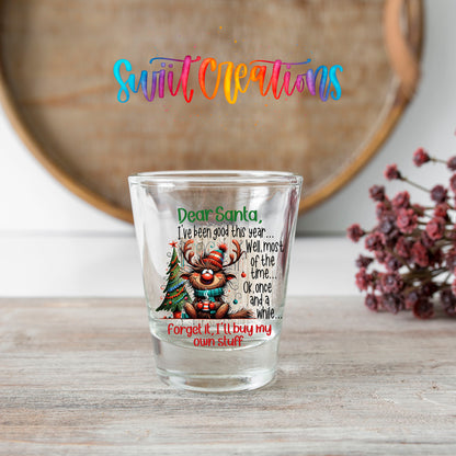 Santa I've Been Good Christmas shot glass transfer on a clear glass beside a wooden barrel and dried flowers
