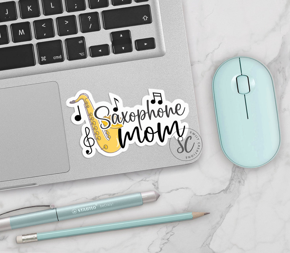 Saxophone Mom vinyl sticker on a silver laptop next to a mint mouse and pens