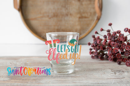 Let's Get Elfed Up Christmas elf shot glass UV DTF transfer shown on a clear shot glass beside dried flowers and a round wooden tray.