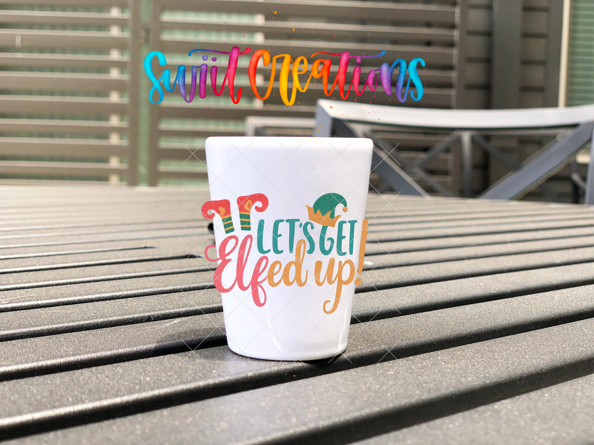 Let's Get Elfed Up Christmas elf shot glass UV DTF transfer shown on a white shot glass on an outdoor slatted table.