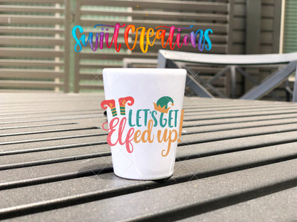 Let's Get Elfed Up Christmas elf shot glass UV DTF transfer shown on a white shot glass on an outdoor slatted table.