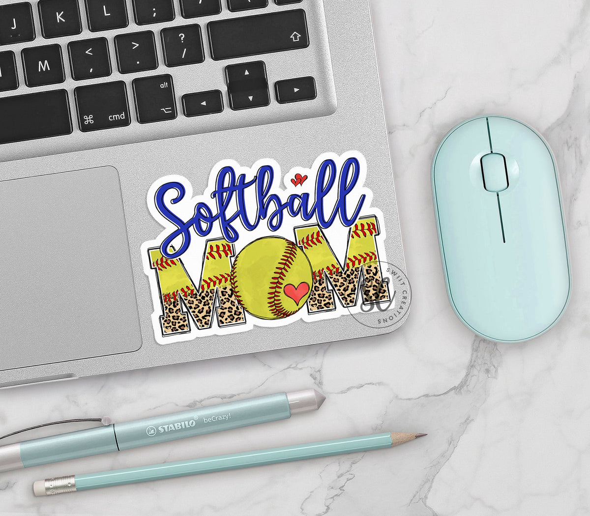 Softball Mom vinyl sticker on a silver laptop next to a mint mouse and pens