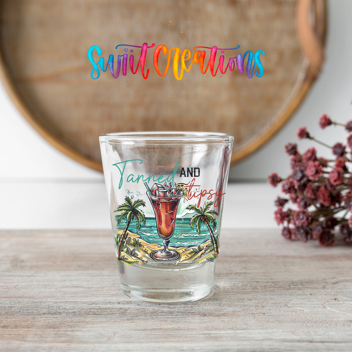 Tanned and Tipsy shot glass transfer on a clear glass beside a wooden barrel and dried flowers