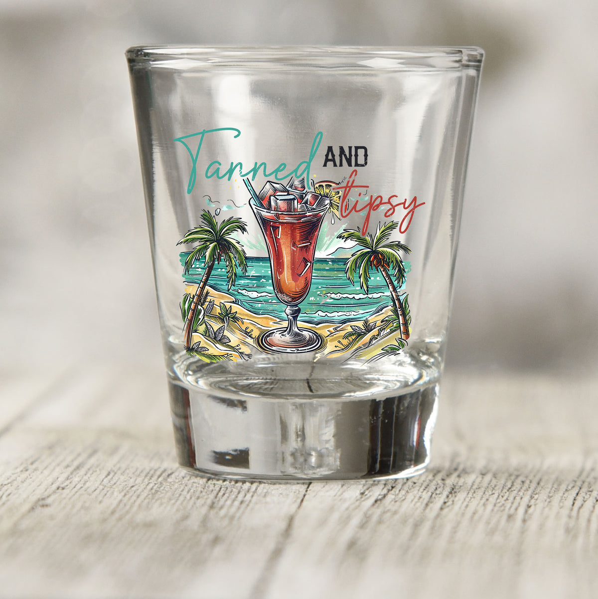 Tanned and Tipsy shot glass UV DTF transfer with cocktail, palm trees, and ocean scene on a clear shot glass
