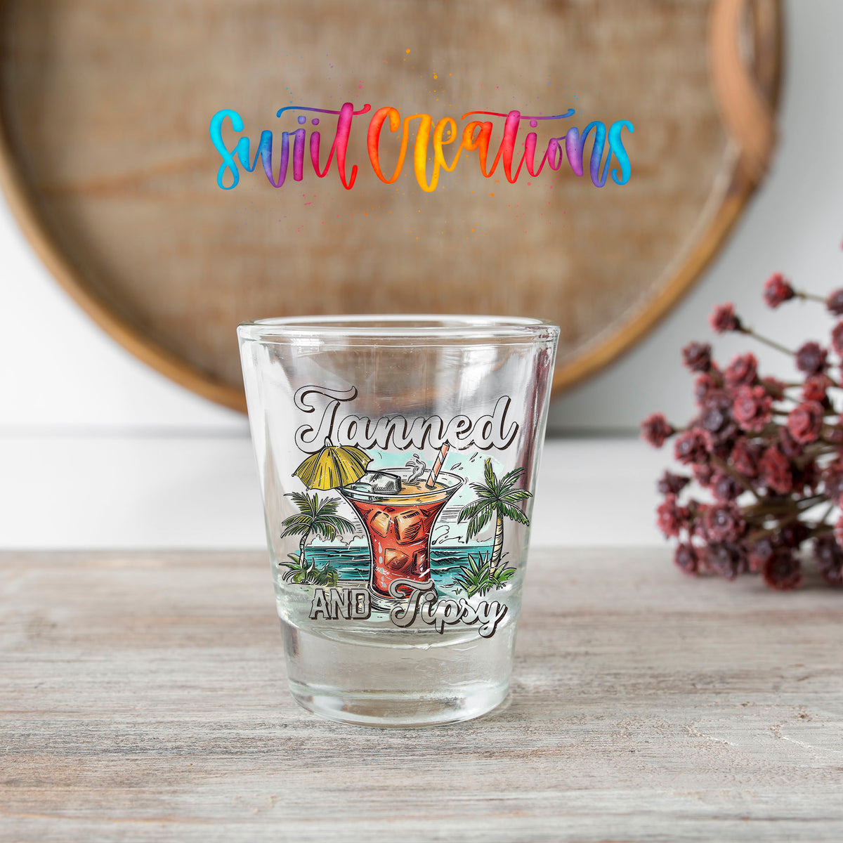 Tanned and Tipsy shot glass transfer on a clear glass beside a wooden barrel and dried flowers