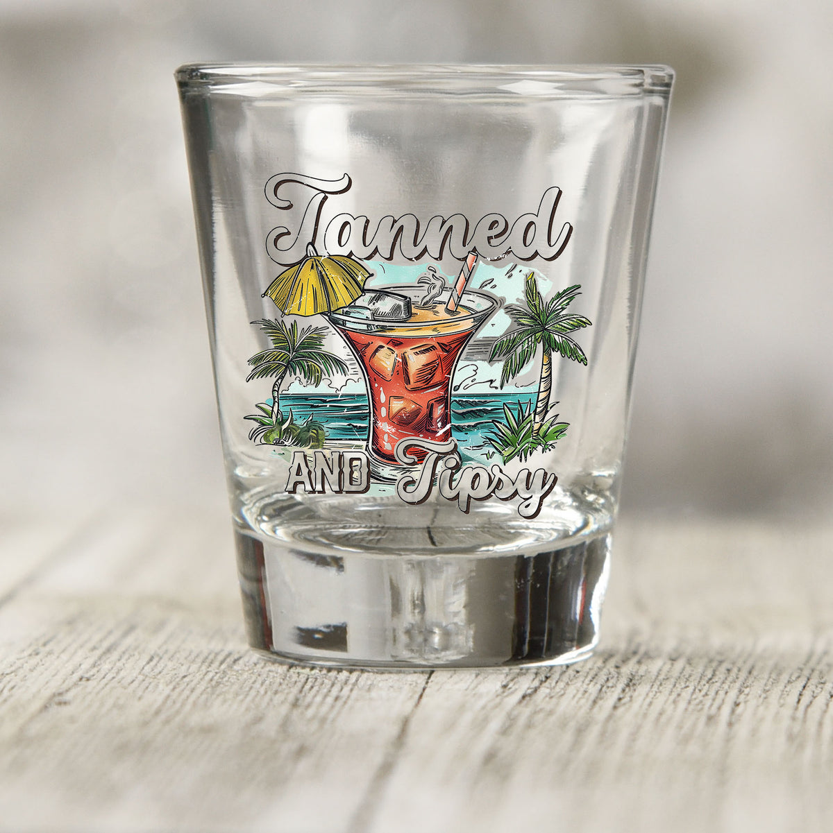 Tanned and Tipsy shot glass UV DTF transfer with umbrella cocktail, palm trees, and ocean scene on a clear shot glass