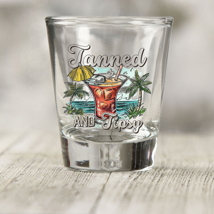 Tanned and Tipsy shot glass UV DTF transfer with umbrella cocktail, palm trees, and ocean scene on a clear shot glass