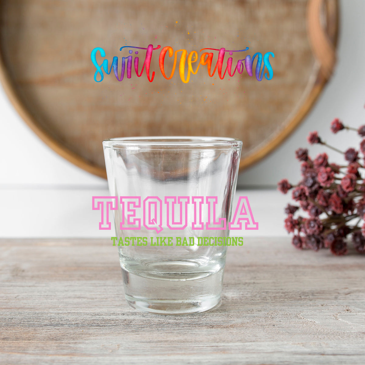 Tequila Tastes Like Bad Decisions shot glass transfer on a clear glass beside a wooden barrel and dried flowers