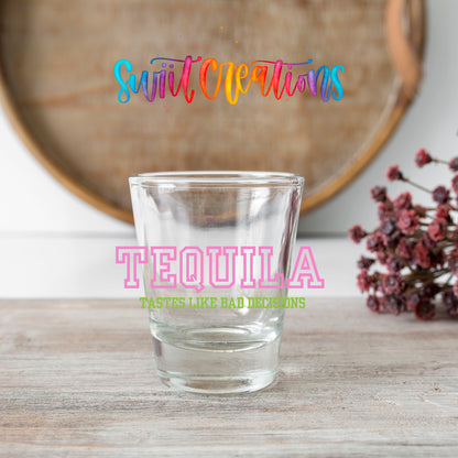 Tequila Tastes Like Bad Decisions shot glass transfer on a clear glass beside a wooden barrel and dried flowers