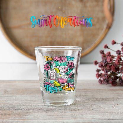 Too Salty For Tears Too Tipsy To Care shot glass transfer on a clear glass beside a wooden barrel and dried flowers